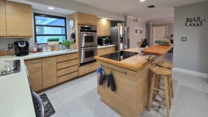 Una cocina moderna con armarios de madera, electrodomésticos de acero inoxidable, una cocina por inducción negra y una pequeña isla con taburetes.