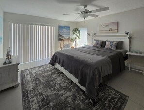Een moderne slaapkamer met een bed, een plafondventilator, een nachtkastje, een wandplank en een raam met jaloezieën.