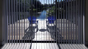 Vista a través de una ventana con persianas verticales, que muestra una terraza de madera con dos sillas azules y una mesa, con vista a un cuerpo de agua y vegetación.