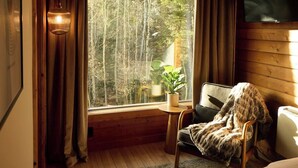 A cozy cabin room with a large window, a chair with a blanket, a small table with a plant, and a bed. - Aspen Hideway · Mountain Modern log Cabin Deck + Hot Tub (Florissant)