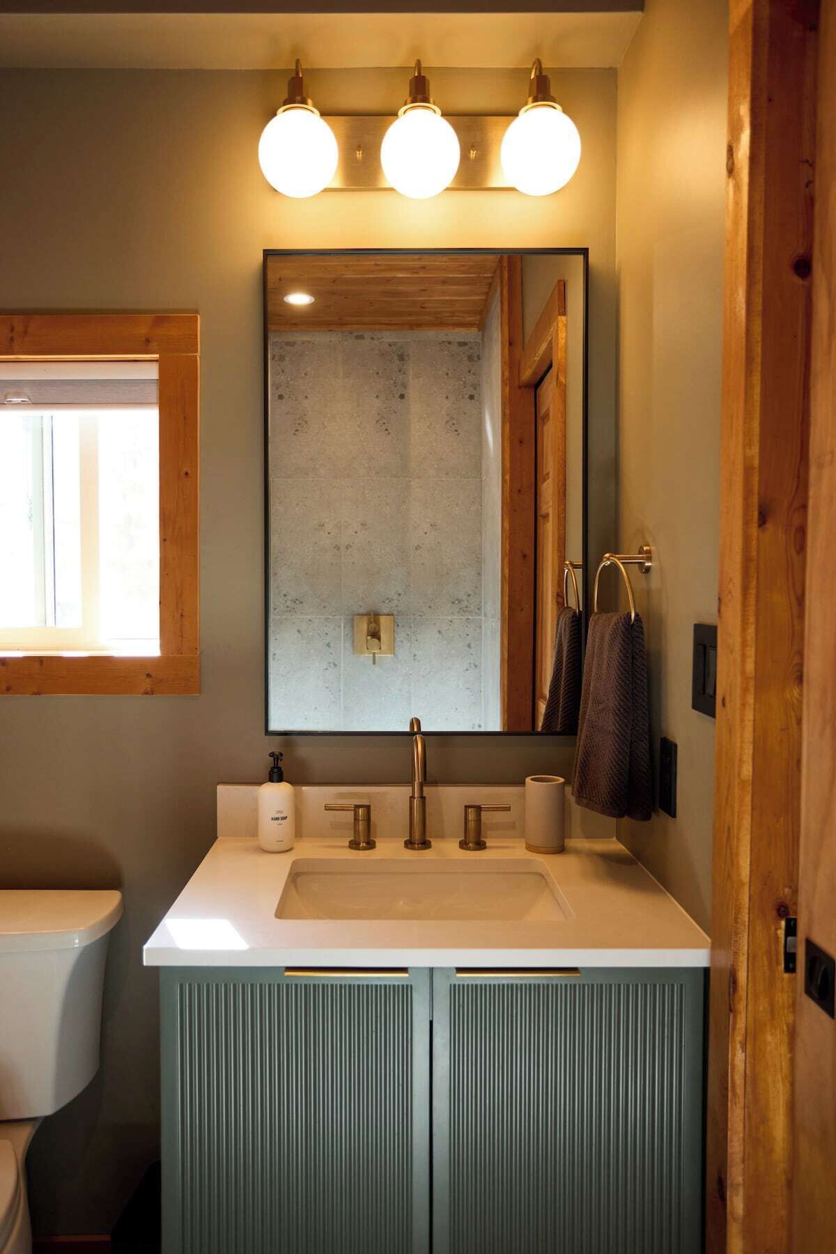 A bathroom with a large mirror, a sink with two faucets, and a toilet. There are three round lights above the mirror and a window to the left.