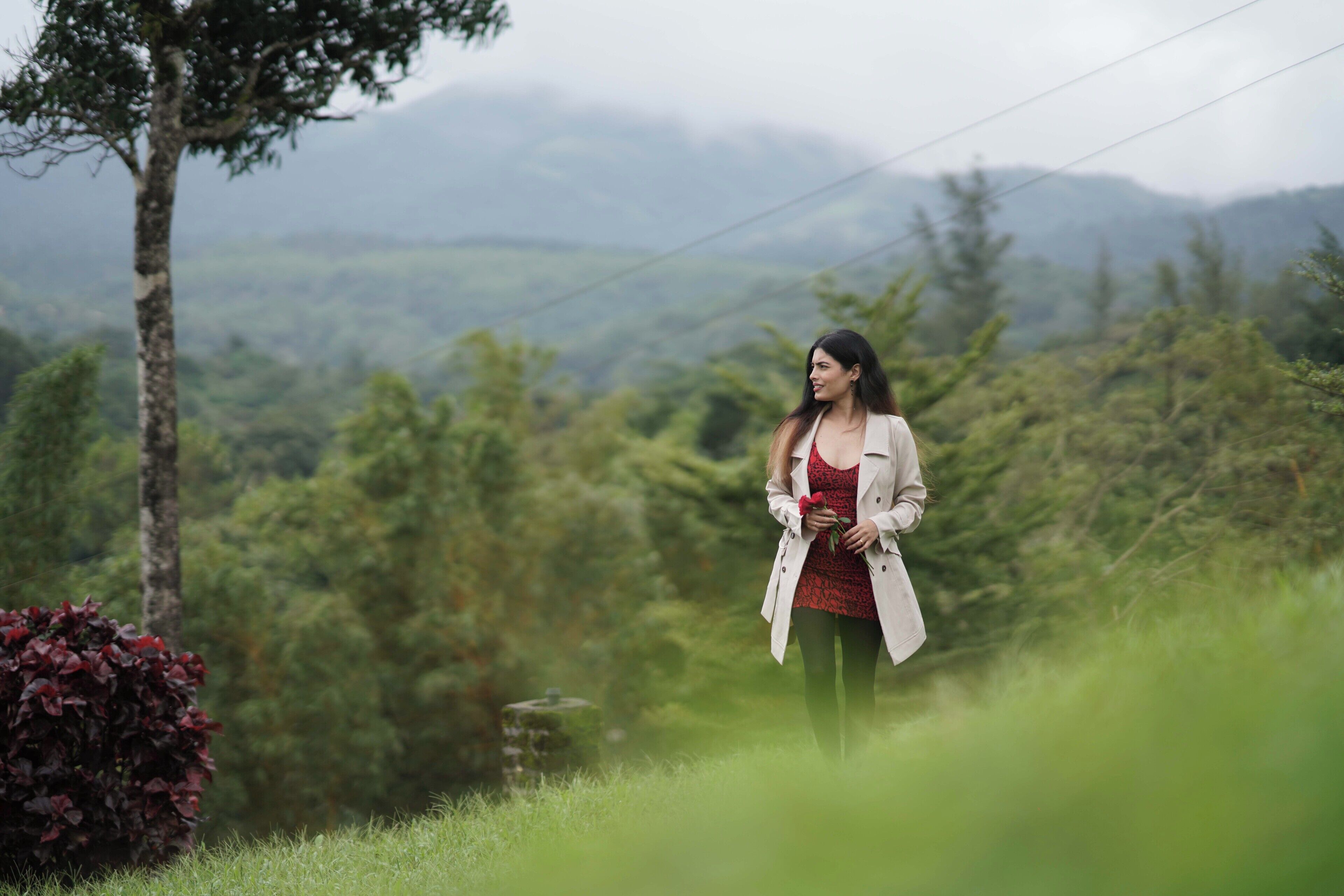 Une femme se tient debout dans un paysage verdoyant, avec des arbres et des collines en arrière-plan.