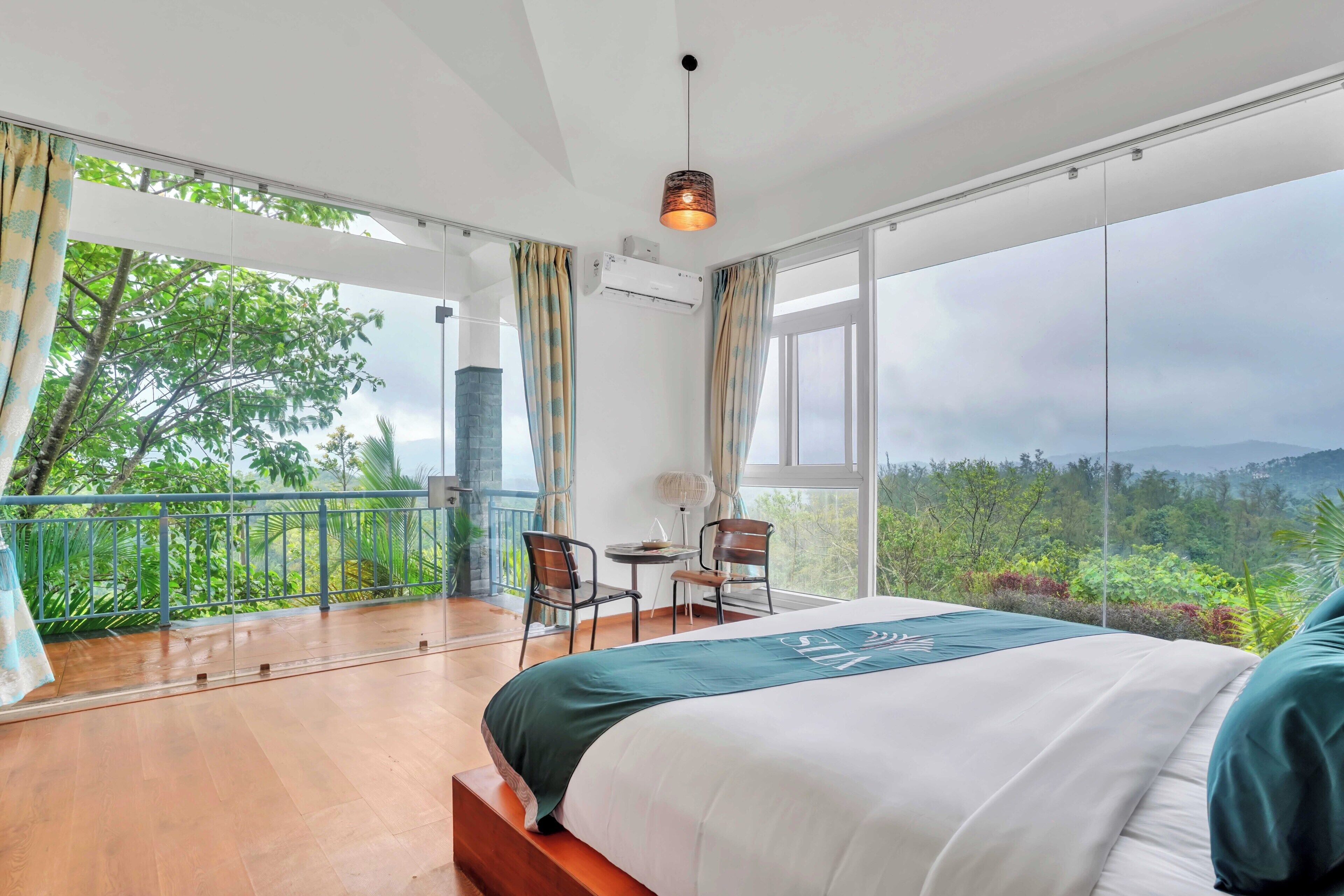 Une chambre avec un grand lit, un balcon donnant sur un espace verdoyant et une table avec des chaises.