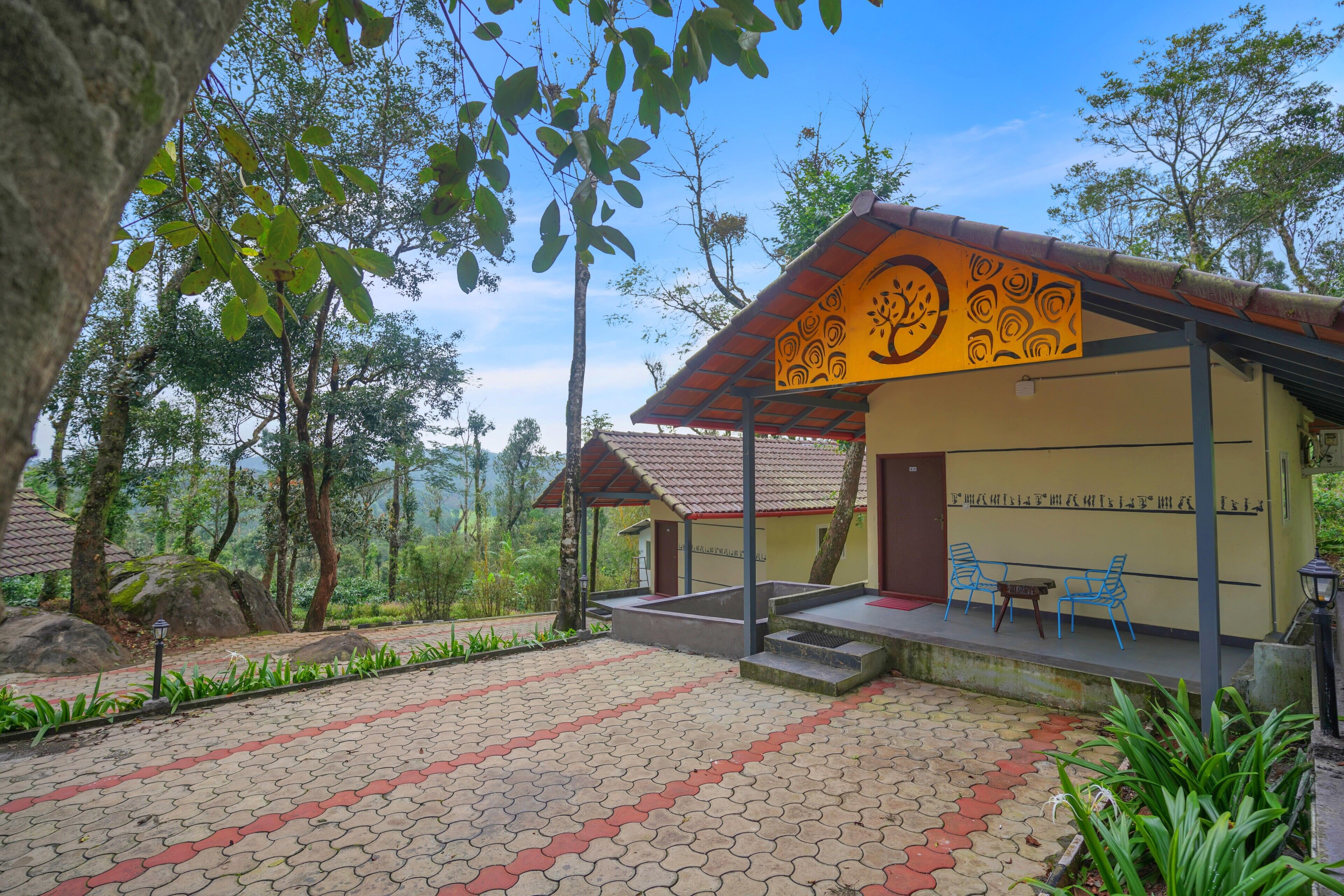 A building with a tiled roof and a yellow wall with black patterns. There are steps leading to a covered area with blue chairs and a table.