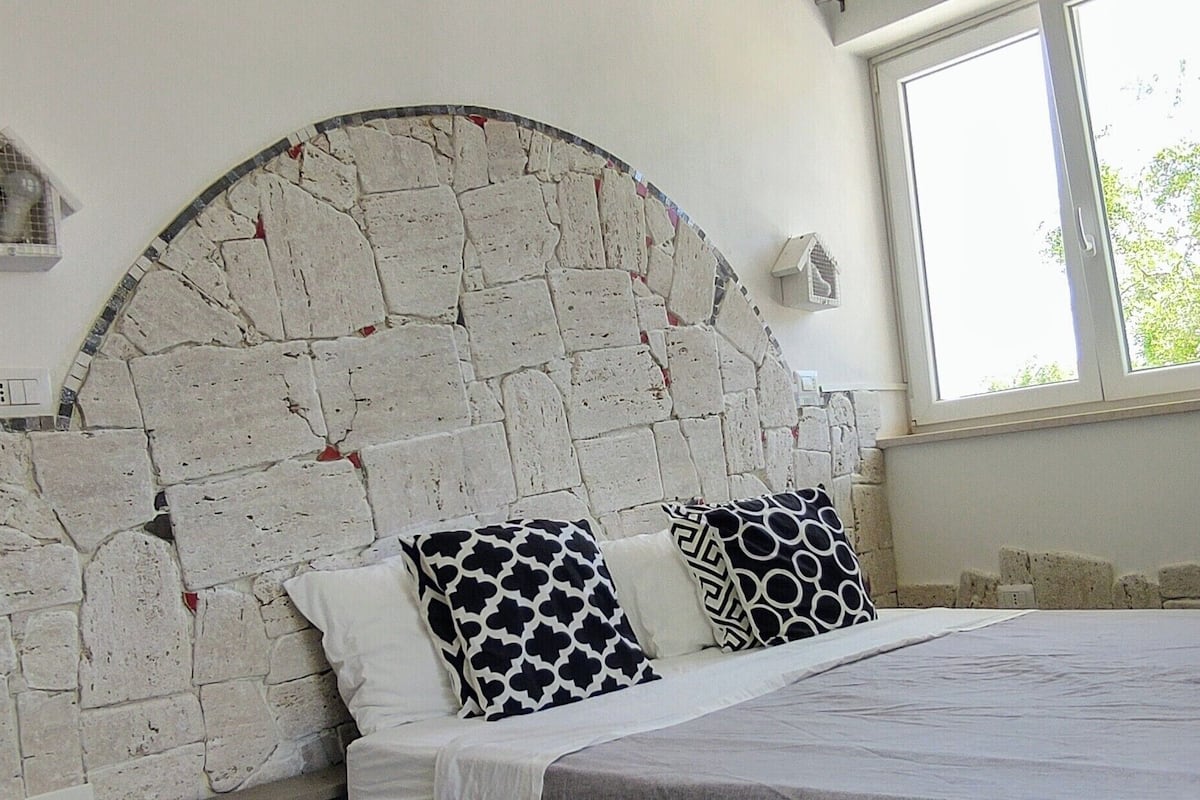 A bedroom with a stone headboard, two patterned pillows, and a window with a view of greenery.