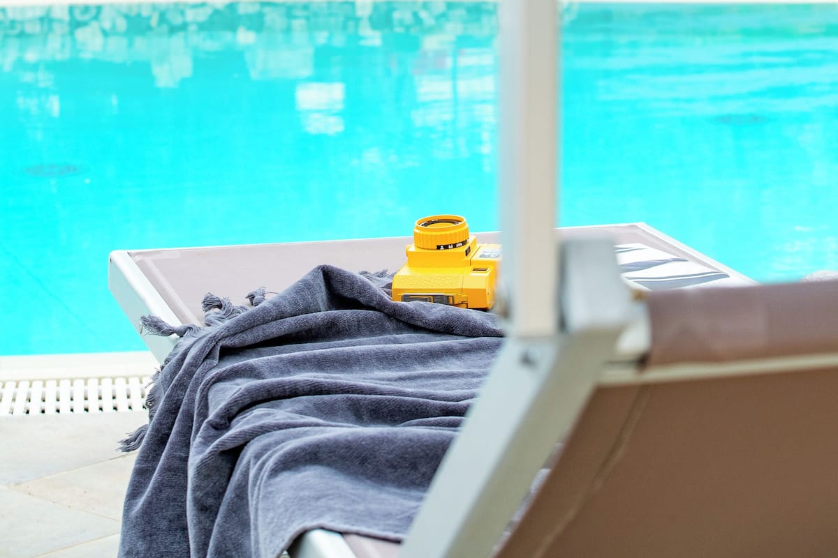 A poolside lounge chair with a towel, a yellow object on the table, and a stone wall with greenery in the background.