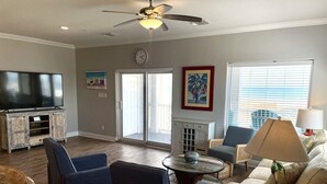 A spacious living room with a flat-screen TV, a ceiling fan, and a large window with blinds. - Beachgate Condo Suites And Hotel 341+342 4 Bedroom Condo (Port Aransas)