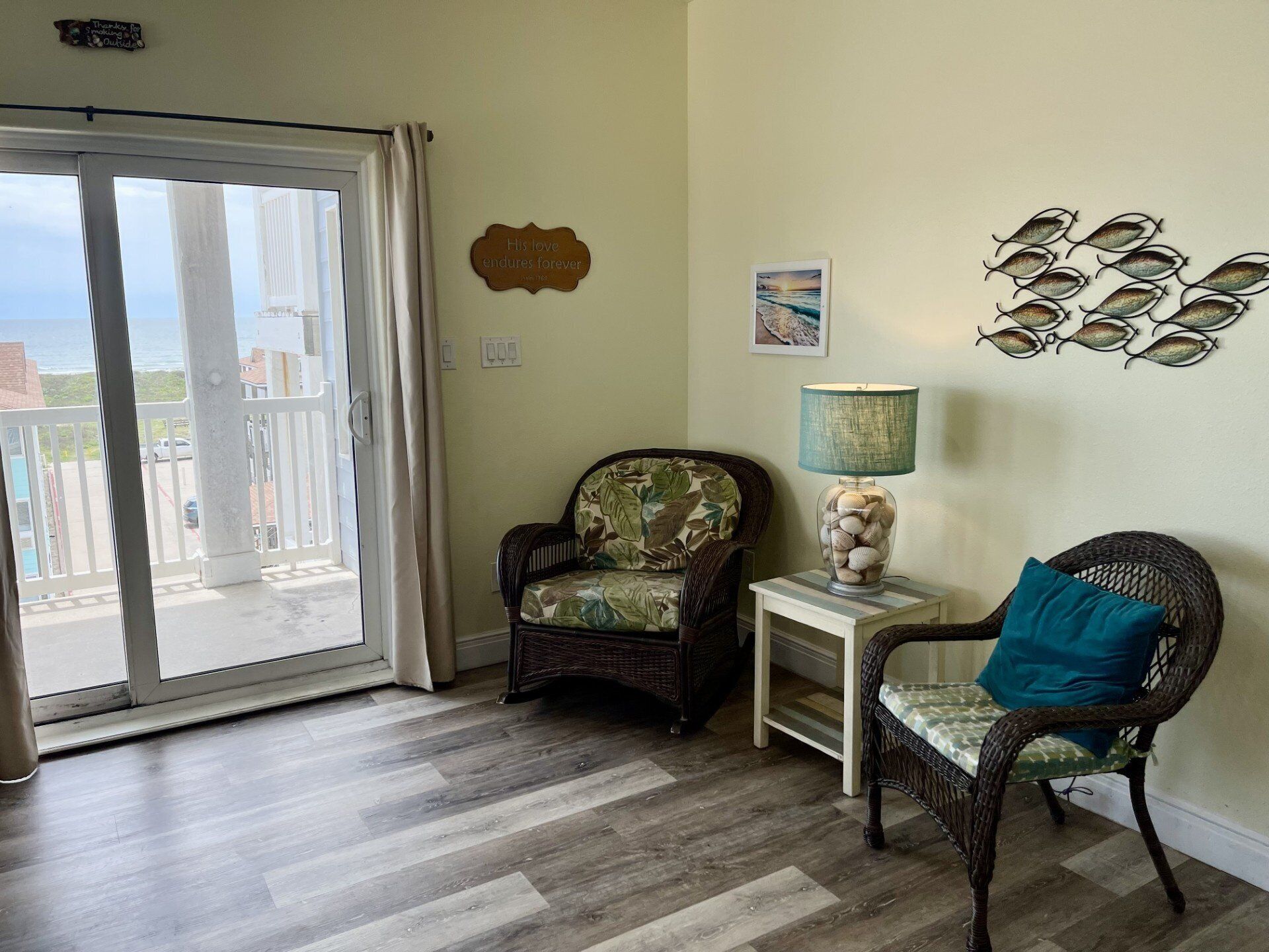 A room with a view to the outside, a wicker chair with a cushion, a small table with a lamp, and a wall with decorative items.