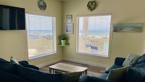 A living room with two couches, a coffee table, and a TV. There are two windows with blinds, a small shelf with a plant, and a painting on the wall. - Beachgate Condo Suites And Hotel 331+332 4 Bedroom Condo (Port Aransas)