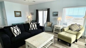 A living room with a dark sofa, a light-colored coffee table, a light armchair, and a small white side table with a lamp. - Beachgate Condo Suites And Hotel 331+332 4 Bedroom Condo (Port Aransas)