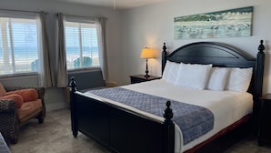 A hotel room with a bed, a chair, a nightstand, a lamp, a ceiling fan, and a painting on the wall. - Beachgate Condo Suites And Hotel 237+238+239 3 Bedroom Condo (Port Aransas)