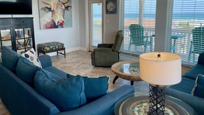 A living room with a blue sofa, a glass coffee table, a green armchair, and a TV on the wall. - Beachgate Condo Suites And Hotel 237+238+239 3 Bedroom Condo (Port Aransas)