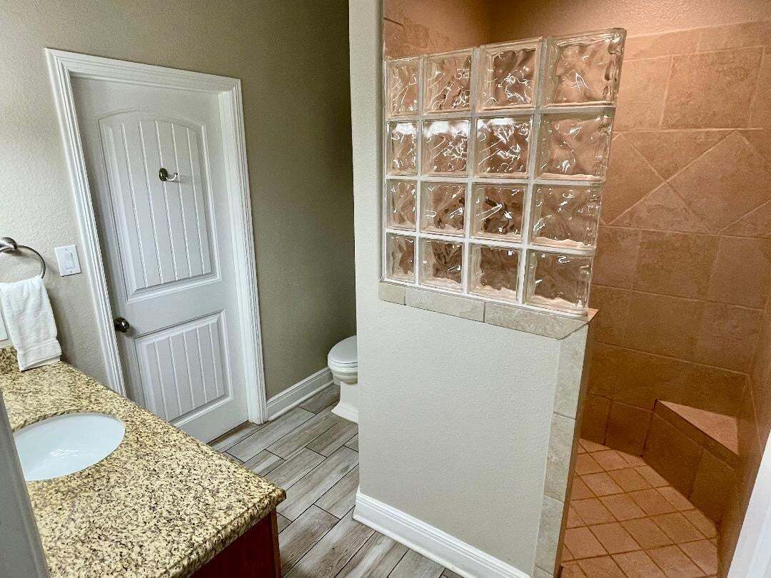 A bathroom with a white door, a sink with a granite countertop, a glass shower enclosure, and a toilet.