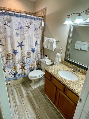 A bathroom with a seashore-themed shower curtain, a toilet, a sink, and a mirror with lights. - Beachgate Condo Suites And Hotel 341 2 Bedroom Condo (Port Aransas)