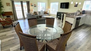 A spacious living room with a round glass table surrounded by wicker chairs, a sofa, and a kitchen area with a dishwasher and a flat-screen TV. - Beachgate Condo Suites And Hotel 322 2 Bedroom Condo (Port Aransas)