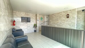 A reception area with a marble wall, a flat-screen TV, a fire extinguisher, a potted plant, a staircase, and a clock. - Pousada Anadia (Anadia)