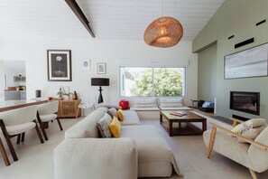 A modern living room with a sofa, chairs, a coffee table, a TV, and a large window with a view of trees. - Villa La Canopée - Ile d'Oléron - Chaucre - 8 pers - swimming pool (Saint-Georges-d'Oléron)
