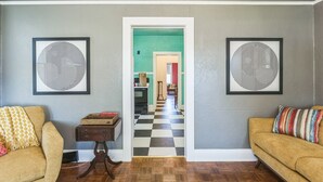 Un pasillo con piso a cuadros que conduce a una zona de cocina. Hay dos cuadros abstractos enmarcados en las paredes, una pequeña mesa de madera con una caja y dos sofás con cojines estampados.