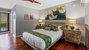 Un dormitorio con una cama grande, cabecera de madera, dos mesitas de noche con lámparas, ventilador de techo y una obra de arte en la pared.