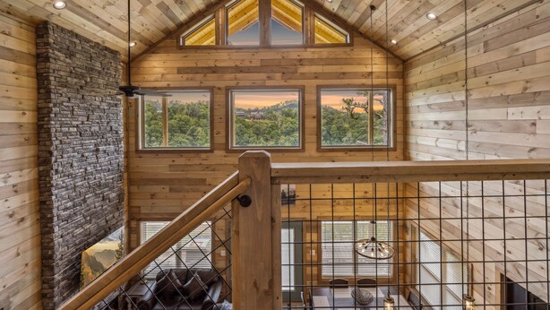 A wooden interior with a stone fireplace, a staircase, and large windows offering a view of the outdoors.