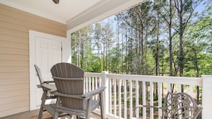 A covered porch with two Adirondack chairs, a white door, and a view of a forest. - River Crossing #1134 2 Bedroom Condo (North Myrtle Beach)