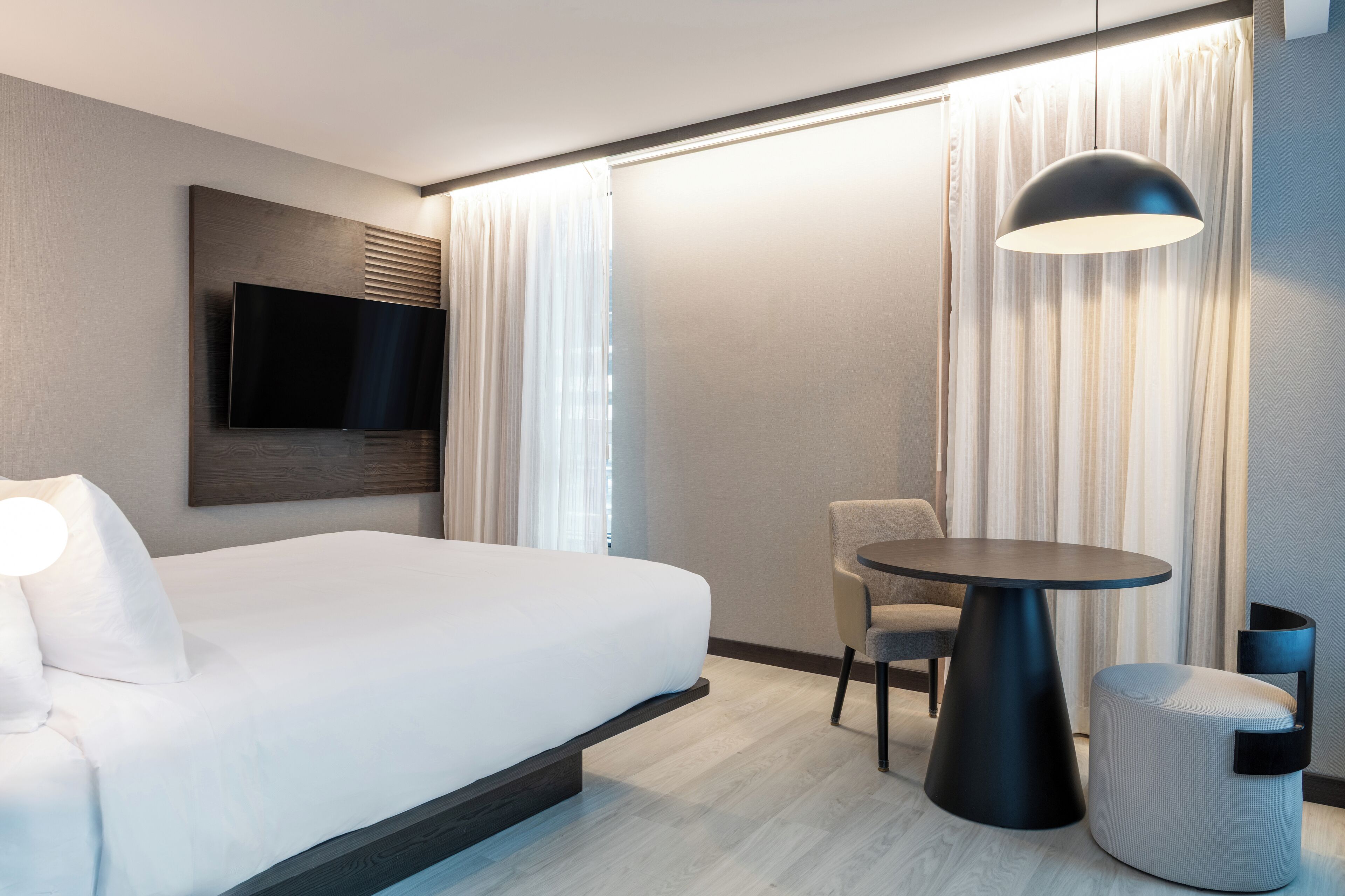 A modern hotel room with a large bed, a flat-screen TV mounted on the wall, a round table, a chair, and a small ottoman.