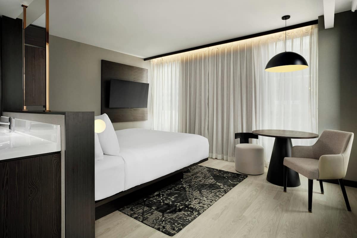 Ein modernes Hotelzimmer mit Bett, Nachttisch, Sessel und einem an der Wand befestigten Fernseher.