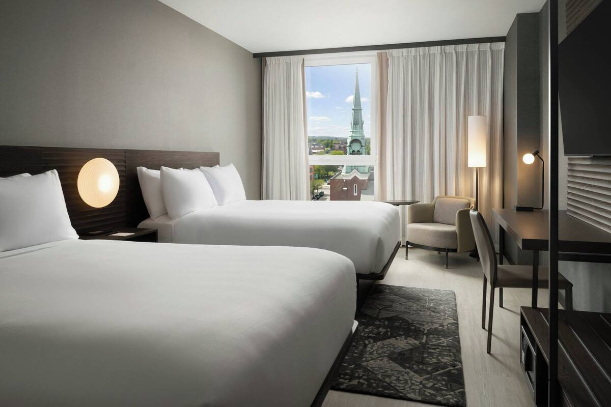 Ein Hotelzimmer mit zwei Betten, einem Schreibtisch, einem Stuhl und Blick auf den Turm einer Kirche durch große Fenster.