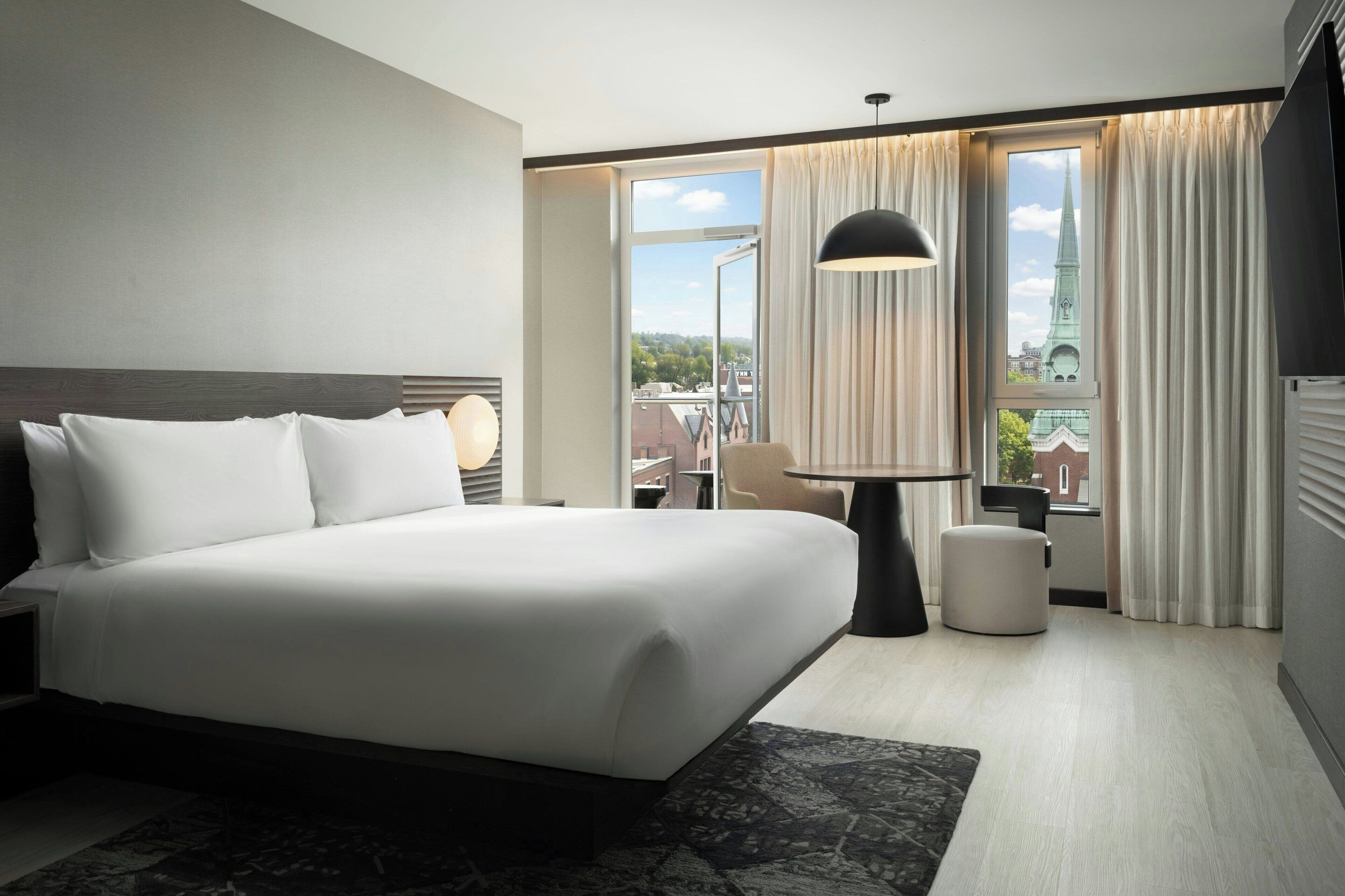 A modern hotel room with a large bed, a desk, a chair, and a view of a cityscape through large windows.