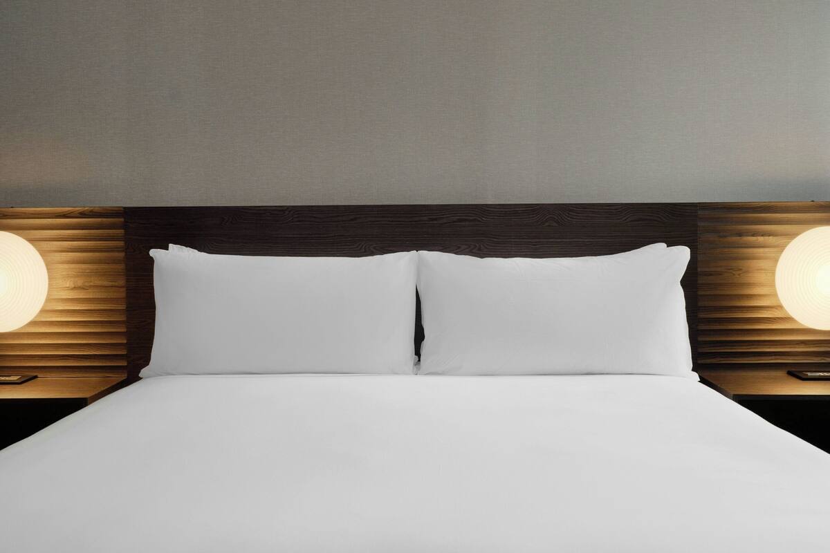 Ein ordentlich bezogenes Bett mit weißen Laken, eingerahmt von zwei Nachttischen mit Holz und jeweils einer Nachttischlampe.