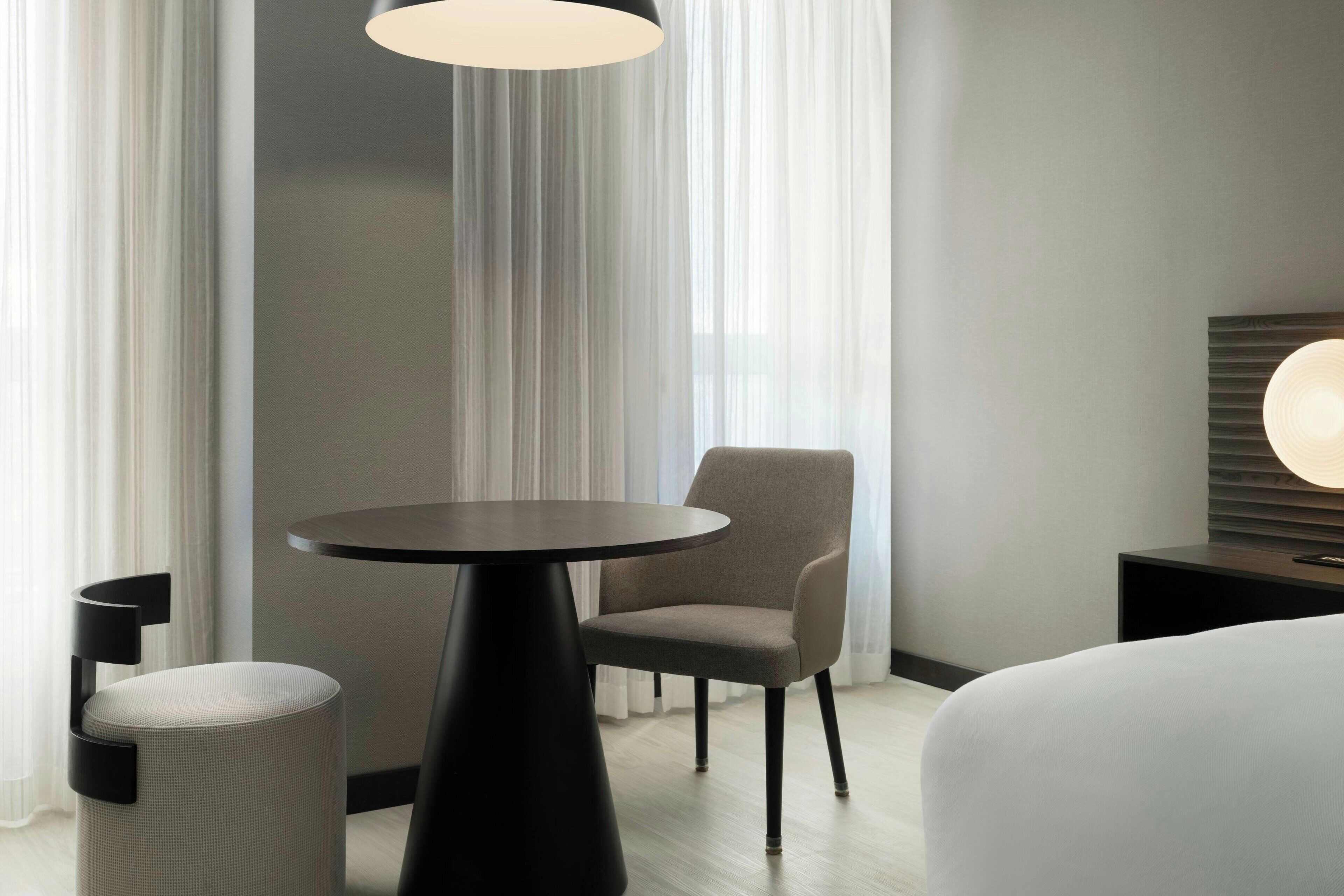 Ein modernes Hotelzimmer mit rundem Tisch, einem gepolsterten Sessel, einem Bett und einem Nachttisch mit Lampe.