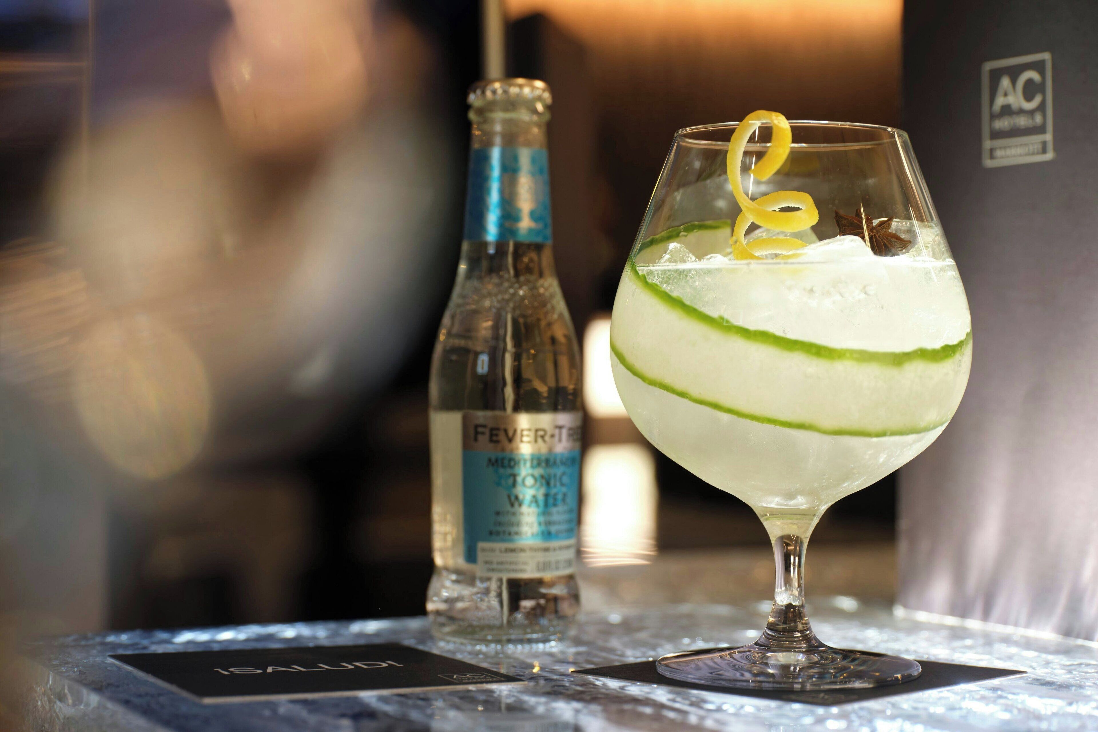 A glass of a clear, layered beverage with a lemon twist garnish, a bottle of Fever-Tree Tonic Water, and a blurred background suggesting a bar or restaurant setting.