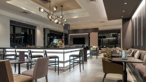 Un hall d’hôtel moderne doté d’un bar central, de plusieurs espaces de détente et d’un grand écran de télévision.