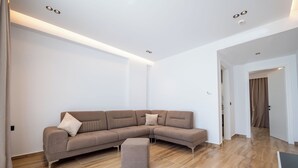 A modern living room with a sectional sofa, a small ottoman, and recessed ceiling lighting. - Alpaso Hotel (Sarande)