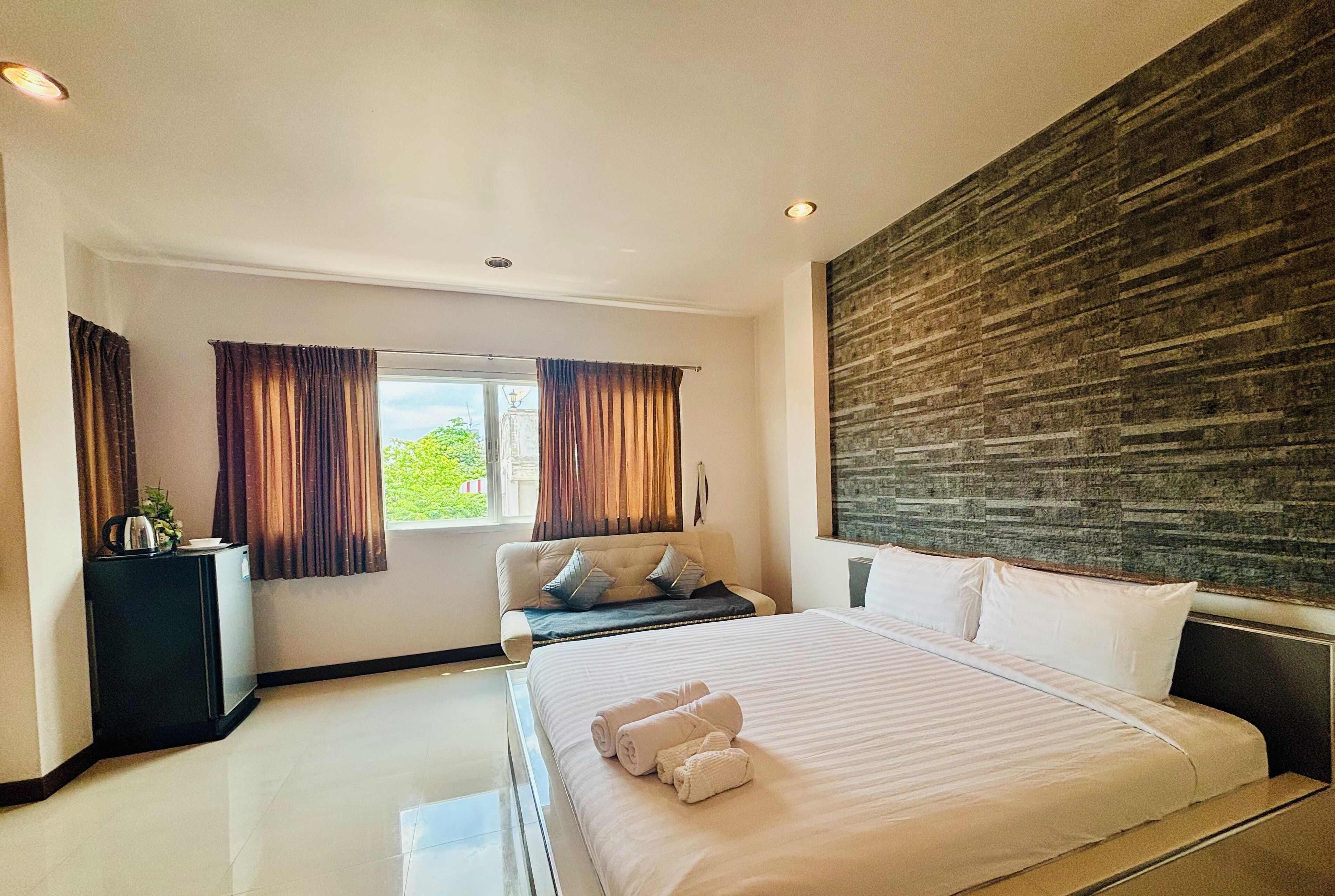 Habitación de hotel moderna con una cama grande, un área de descanso con sofá y almohadas, una mesita con una planta y vistas a zonas verdes.