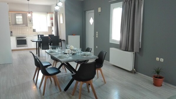 A modern dining area with a rectangular table set for four, surrounded by black chairs with wooden legs. A potted plant is visible in the corner.