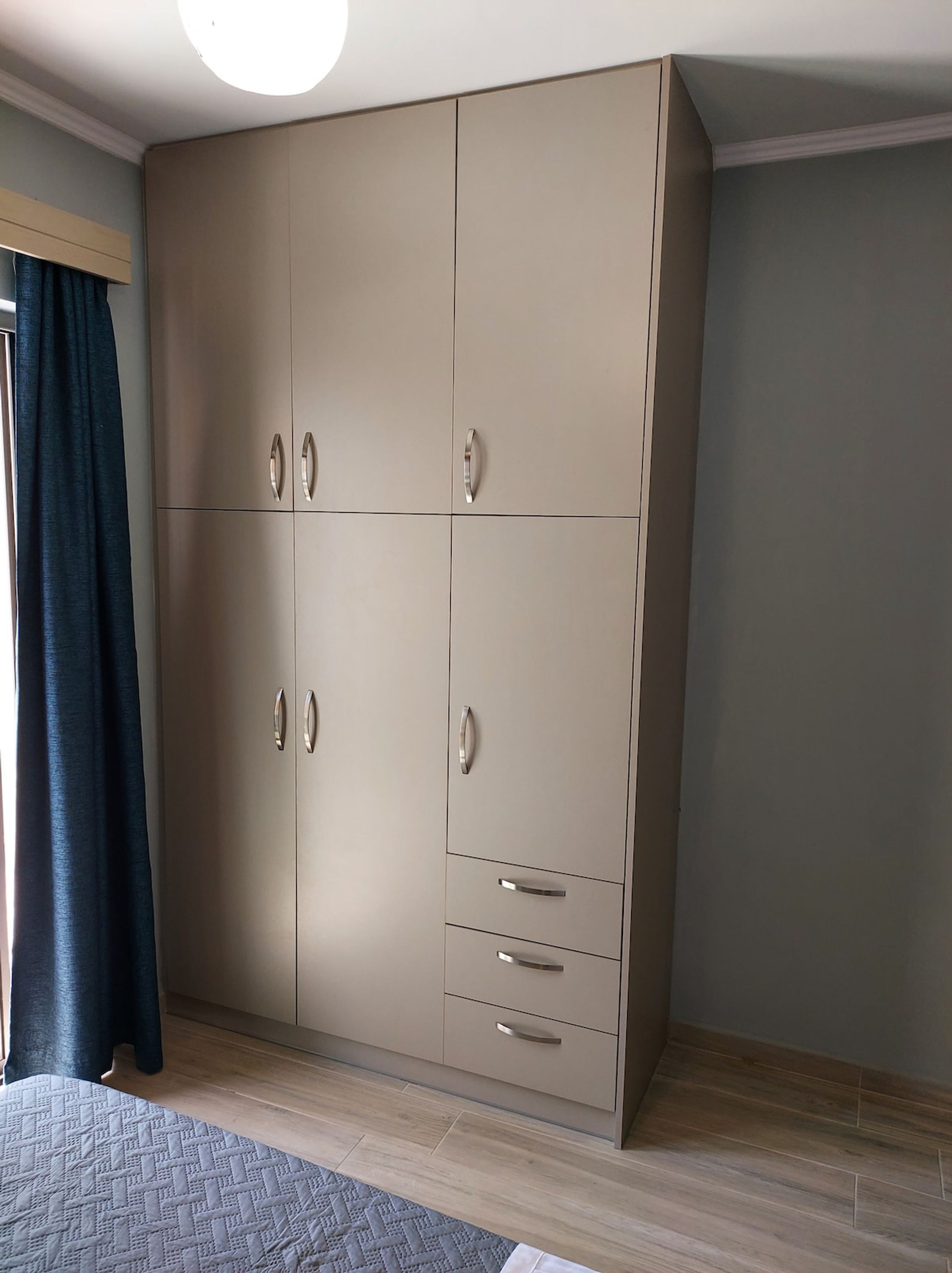 A room with a large wardrobe, a bed with a patterned bedspread, and a window with curtains.