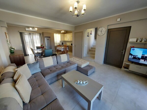 A modern living room with a sectional sofa, a coffee table, and a television. There is a kitchen in the background with a dining area.