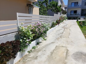 A pathway flanked by a low white fence with flowering plants, leading to a modern building with balconies and a clear sky. - Rethymno Suburb Maisonette (Rethymno)