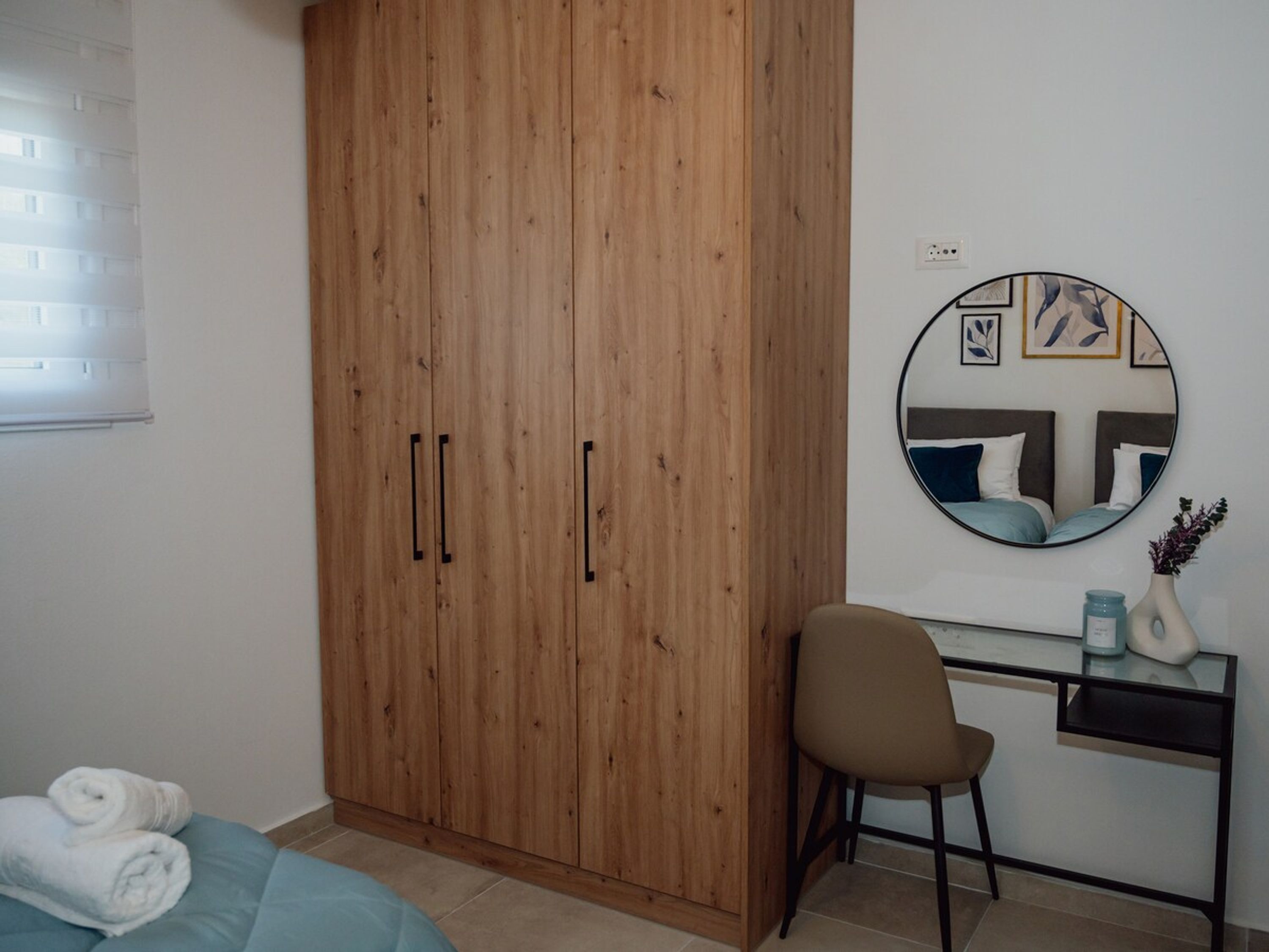 Una camera da letto con un armadio in legno, uno specchio rotondo, una scrivania con una sedia e un letto con asciugamani arrotolati.