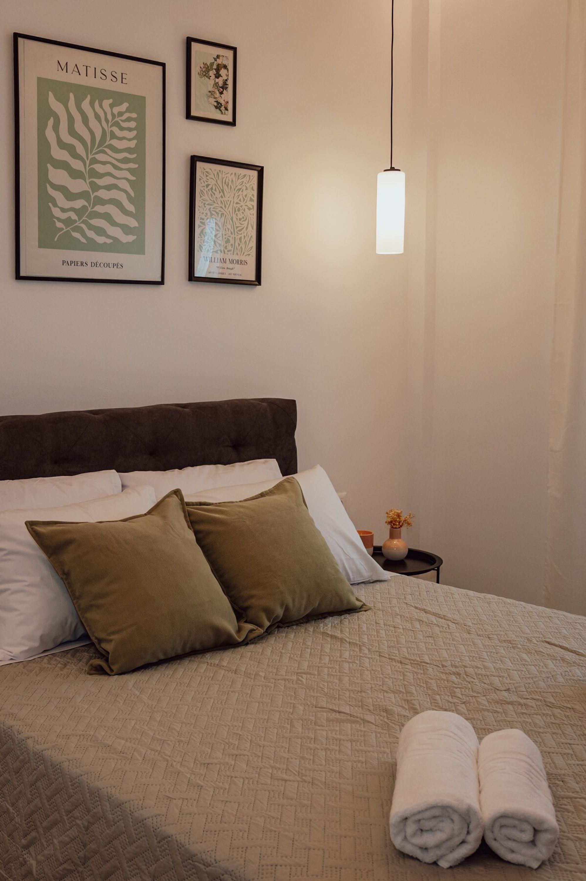 A neatly made bed with a quilted cover, two pillows, and rolled towels on a nightstand.