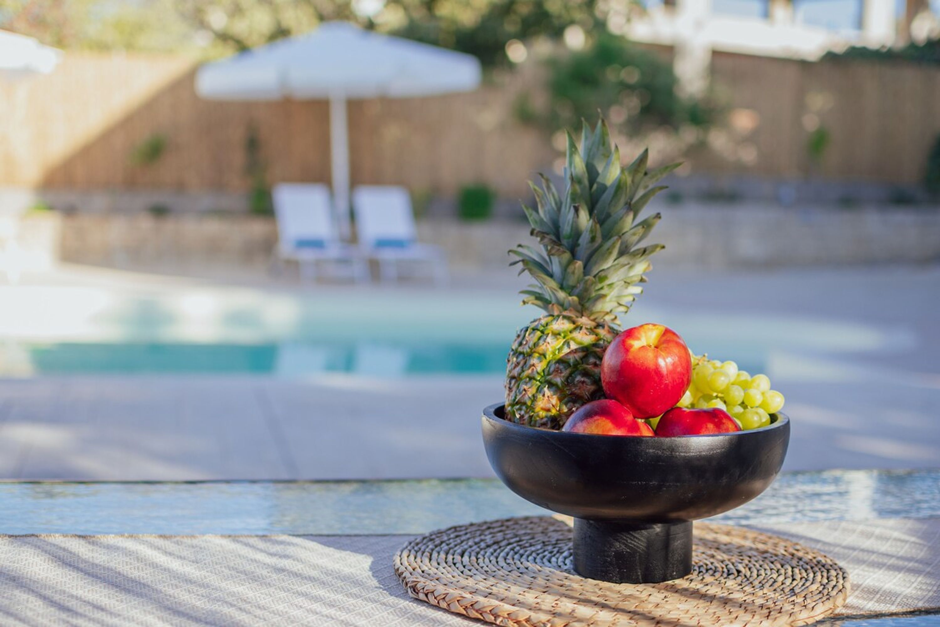 Un frutero sobre una mesa, con una piscina y sillones de descanso al fondo.