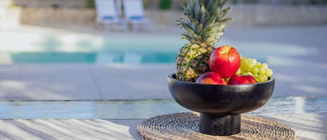 A fruit bowl on a table with a pool and lounge chairs in the background.