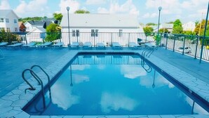 Una piscina rectangular con solárium alicatado, rodeada por una cerca metálica, y un edificio blanco al fondo.