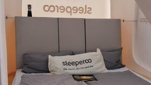 A bed with a grey headboard, a pillow with the text "sleeperoo Die Nacht, der Ort und Du.", and a book on the bed. - Romantik zwischen Weinreben - Cube (Roemerberg)