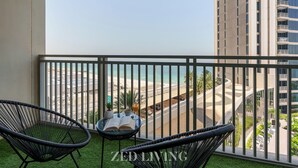 Un balcon avec deux fauteuils en osier noir, une petite table sur laquelle se trouvent un livre et un verre de jus d’orange, et une vue sur une plage et des bâtiments.