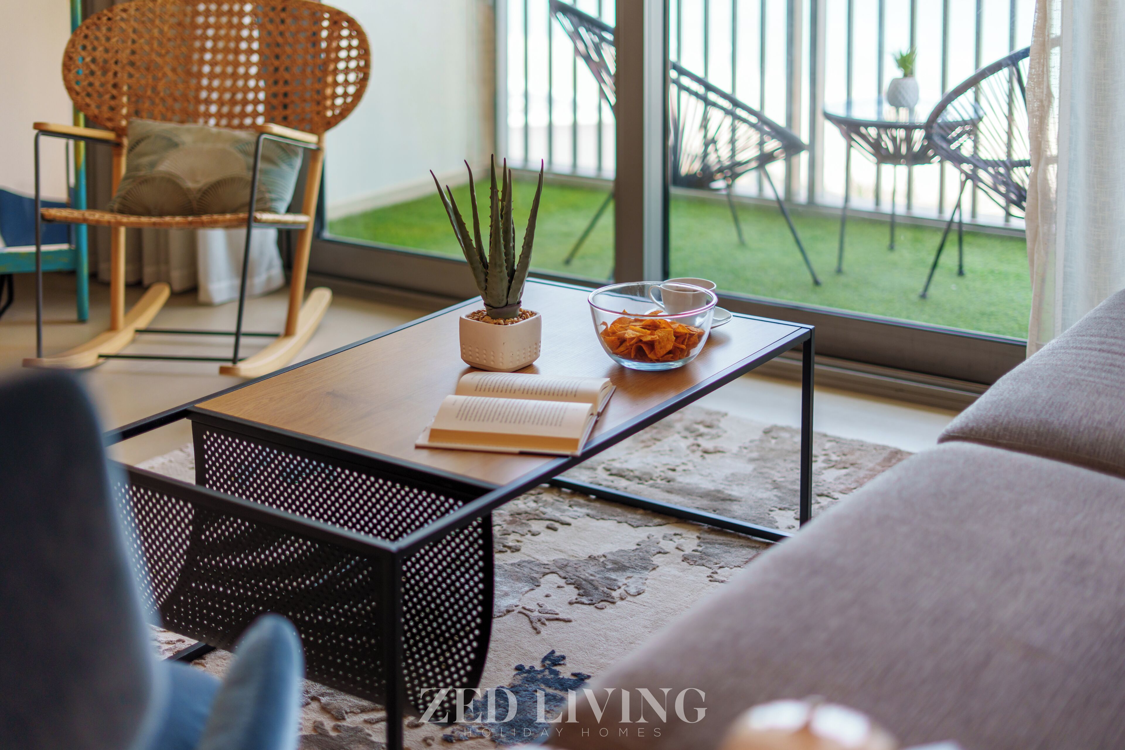 Un salon comprenant un canapé, un fauteuil à bascule, une table basse avec un livre et une plante en pot, et donnant sur un balcon aménagé avec du mobilier d’extérieur.