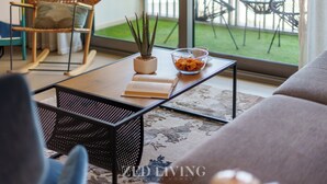 Un salon comprenant un canapé, un fauteuil à bascule, une table basse avec un livre et une plante en pot, et donnant sur un balcon aménagé avec du mobilier d’extérieur.