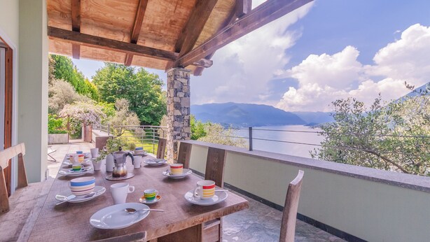 A wooden table set with plates, cups, and a coffee pot on a balcony with a view of mountains and a lake.