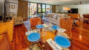 A living room with a sofa, dining table with blue plates, wicker chairs, and a TV. - *Beach* KingBd * Resort * Pool * Adult Hot Tub * (Kihei)